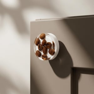 Puede incluir: Un pomo de armario redondo y transparente con granos de café tostados incrustados. El pomo está montado en una puerta de armario de tono neutro, creando una estética minimalista. Los granos de café son de color marrón oscuro.