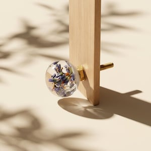 Puede incluir: Un pomo de armario redondo y transparente con flores de lavanda secas en su interior. El pomo está fijado a una superficie de madera clara con un perno dorado. El fondo es de color beige neutro.
