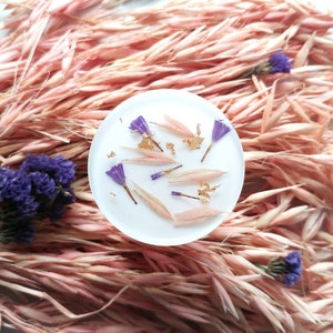May include: A white round coaster with dried flowers and gold leaf embedded in clear resin. The coaster is surrounded by dried pink oat grass.