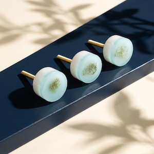 Könnte beinhalten: Drei dekorative Schrankknöpfe mit hellblauem und weißem Design, jeweils mit einem kleinen, getrockneten Pflanzenelement. Die Knöpfe haben goldfarbene Stiele und sind auf einer dunkelblauen Oberfläche angeordnet. Diese Knöpfe sind ideal, um Möbeln eine einzigartige Note zu verleihen.