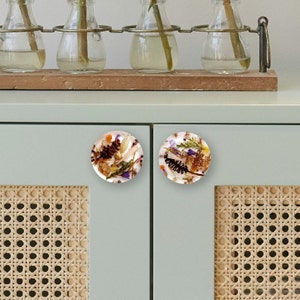 May include: Two round cabinet knobs with a white background and a colourful pressed flower design. The knobs are made of resin and have a glossy finish.