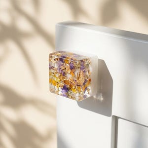 Puede incluir: Un tirador de armario rectangular transparente con flores secas y pan de oro encapsulados en resina. El tirador está montado en una puerta de armario blanca. Las flores son moradas, amarillas y blancas. El pan de oro añade un toque de elegancia.