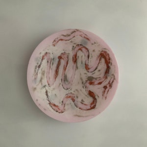 May include: A round, pink ceramic bowl with a white and brown marbled design. The bowl has a smooth, glossy finish.