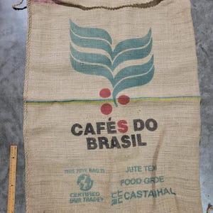 May include: A large, beige burlap sack with the text "CAFÉS DO BRASIL" in black and red. The sack features a teal and red graphic of coffee plant elements. The words "THIS JUTE BAG IS" and "FOOD GRADE" are also visible.