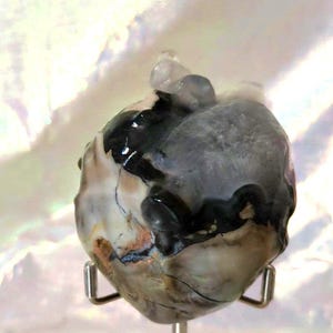 May include: A heart-shaped stone with a unique, swirling pattern of black, white, and gray. The stone is on a white stand with a silver metal base.