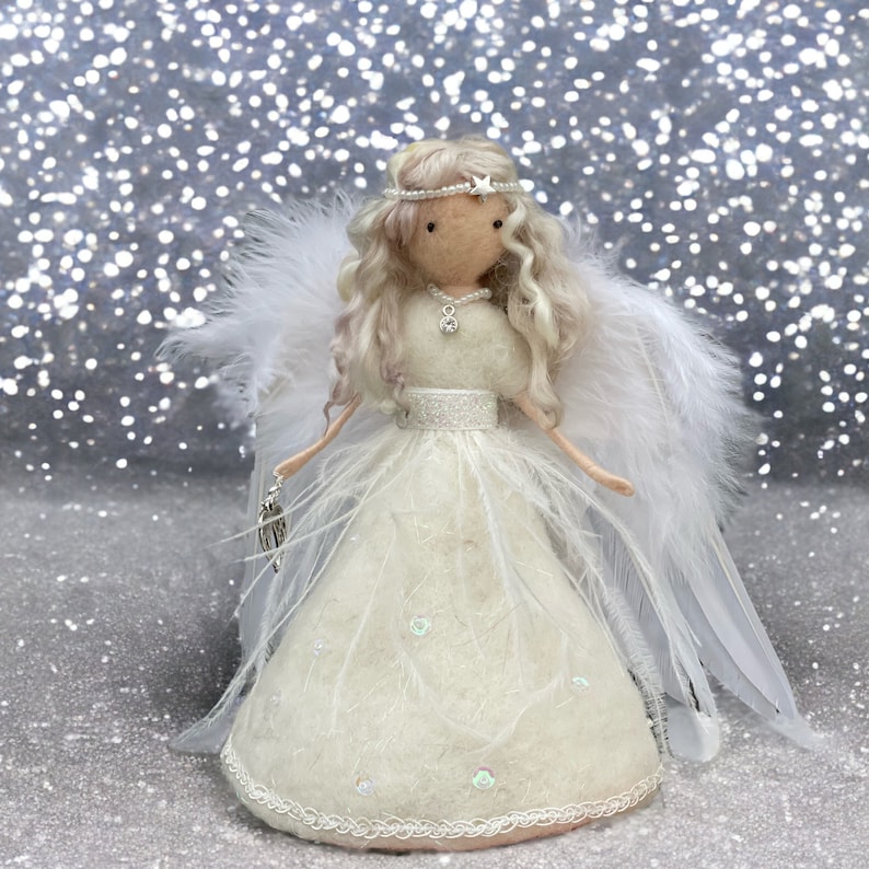 May include: A handcrafted angel doll with a cream-colored dress adorned with feathers and sequins. The doll has long, wavy blonde hair, a pearl and star headband, and large white feathered wings. It holds a silver charm and is set against a sparkly background.