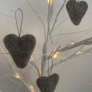 May include: Three grey felt heart decorations suspended from a white decorative tree with small lights. The hearts have a textured appearance and are hung by silver-coloured strings, creating a simple, yet elegant display. The tree's branches are bare, and the background is a soft, neutral tone.