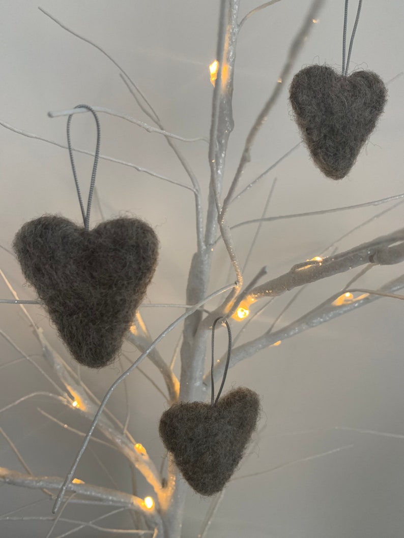May include: Three grey felt heart decorations suspended from a white decorative tree with small lights. The hearts have a textured appearance and are hung by silver-coloured strings, creating a simple, yet elegant display. The tree's branches are bare, and the background is a soft, neutral tone.