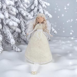May include: A handcrafted angel doll with a cream-colored dress, feathered wings, and a pearl-adorned headband. The doll has long, blonde hair and is set against a snowy backdrop with a snow-covered evergreen tree. The angel is a decorative item.