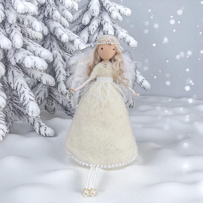 May include: A handcrafted angel doll with a cream-colored dress, feathered wings, and a pearl-adorned headband. The doll has long, blonde hair and is set against a snowy backdrop with a snow-covered evergreen tree. The angel is a decorative item.