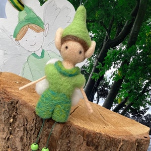 May include: A handcrafted felt fairy doll, dressed in green with a matching pointed hat and small white wings. The fairy is displayed on a wooden surface, with a watercolor illustration of a fairy in the background. The doll has green beads hanging from its legs.