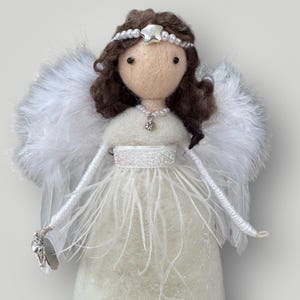 May include: A handcrafted angel doll with a cream-colored felt body and fluffy white feather wings. The doll has brown hair, a pearl and star headband, and a pearl necklace with a pendant. The angel's dress is adorned with white feathers and a sparkly belt.