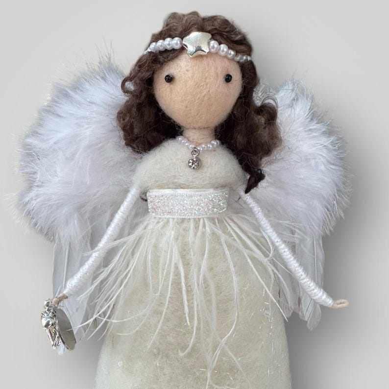 May include: A handcrafted angel doll with a cream-colored felt body and fluffy white feather wings. The doll has brown hair, a pearl and star headband, and a pearl necklace with a pendant. The angel's dress is adorned with white feathers and a sparkly belt.