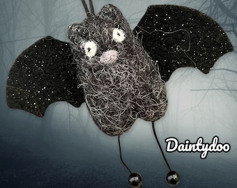 May include: A black felt bat with glitter wings and a white face. The bat has long black legs with black beads at the ends. The bat is hanging from a black string. The text "Dainytdoo" is in the bottom right corner of the image.