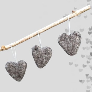 May include: Three grey, heart-shaped decorations suspended from a light brown branch. Each heart hangs by a thin, white string. The hearts have a textured, felt-like appearance, set against a soft grey background.