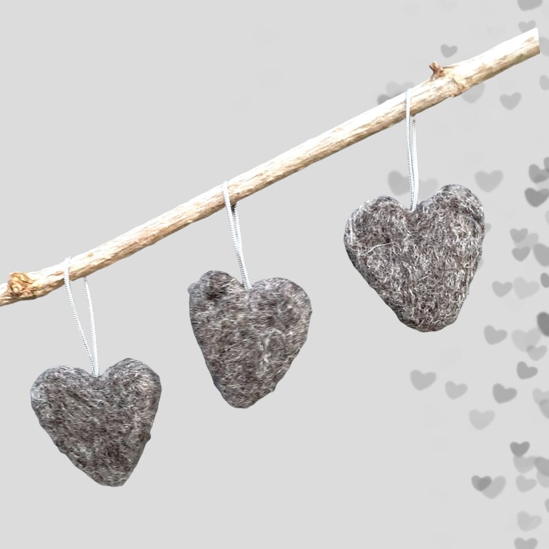 May include: Three grey, heart-shaped decorations suspended from a light brown branch. Each heart hangs by a thin, white string. The hearts have a textured, felt-like appearance, set against a soft grey background.