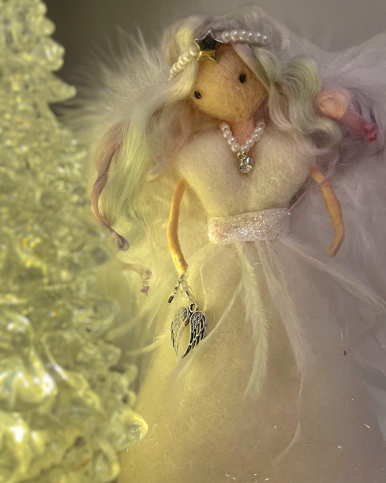 May include: A handmade angel figurine with a white dress and feathered wings. The angel has long, flowing hair with pastel highlights and wears a pearl necklace and a star-shaped crown. It holds a charm with silver wings.