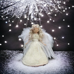 May include: A handcrafted angel doll with blonde hair, a white dress, and feathered wings. The doll has a silver halo and holds a small lantern. The background features twinkling lights and a snowy surface, creating a festive holiday scene.