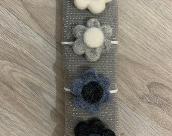 May include: Four felt flower hair ties in different colors. The hair ties are on a gray background. The flowers are white with gray centers, white with gray centers, blue with black centers, and black with blue centers.