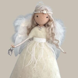 May include: A handcrafted angel doll with a cream-colored dress adorned with feathers and sequins. The doll has long, wavy, light-colored hair, a pearl and star headband, and a pearl necklace. It holds a silver charm and has feathered wings.