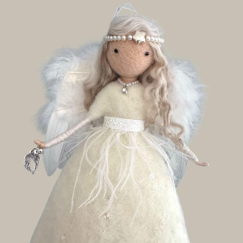 May include: A handcrafted angel doll with a cream-colored dress adorned with feathers and sequins. The doll has long, wavy, light-colored hair, a pearl and star headband, and a pearl necklace. It holds a silver charm and has feathered wings.
