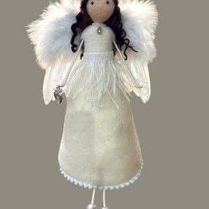 May include: A handmade angel doll with a cream-colored felt dress, white feather wings, and a beaded halo. The doll has brown curly hair and holds a silver charm. The dress is adorned with white feathers and a beaded trim. The doll's legs end in two silver beads.