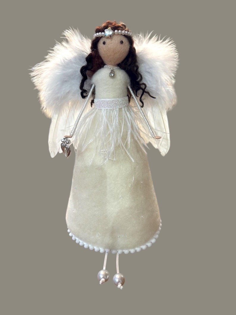 May include: A handmade angel doll with a cream-colored felt dress, white feather wings, and a beaded halo. The doll has brown curly hair and holds a silver charm. The dress is adorned with white feathers and a beaded trim. The doll's legs end in two silver beads.