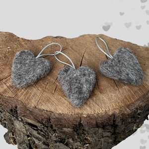 May include: Three grey felt heart decorations with silver string loops. The hearts are presented on a rustic wooden surface, ideal for Valentine's Day or other celebrations. Each heart is approximately 7.6 cm wide.