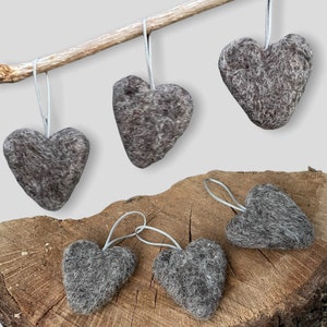 May include: Five grey felt heart decorations with silver hanging ribbons. The hearts are displayed on a wooden surface and a small branch. The ornaments have a textured, woolly appearance, and are in various sizes and orientations.