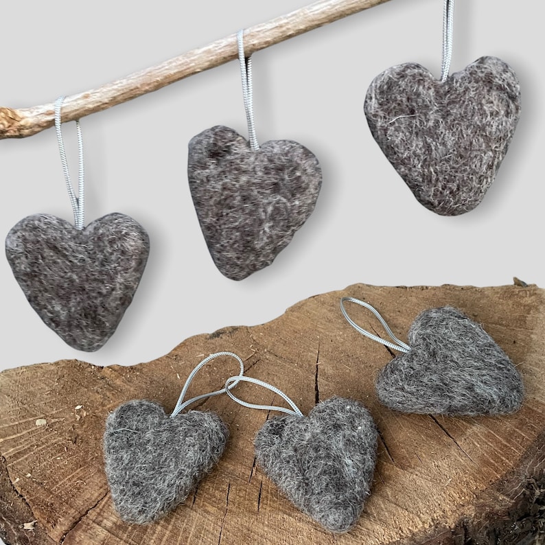 May include: Five grey felt heart decorations with silver hanging ribbons. The hearts are displayed on a wooden surface and a small branch. The ornaments have a textured, woolly appearance, and are in various sizes and orientations.