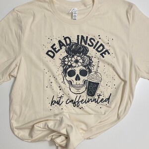 May include: A beige t-shirt with a black graphic design of a skull with flowers in its hair holding a iced coffee. The text "Dead Inside but Caffeinated" is printed below the skull.