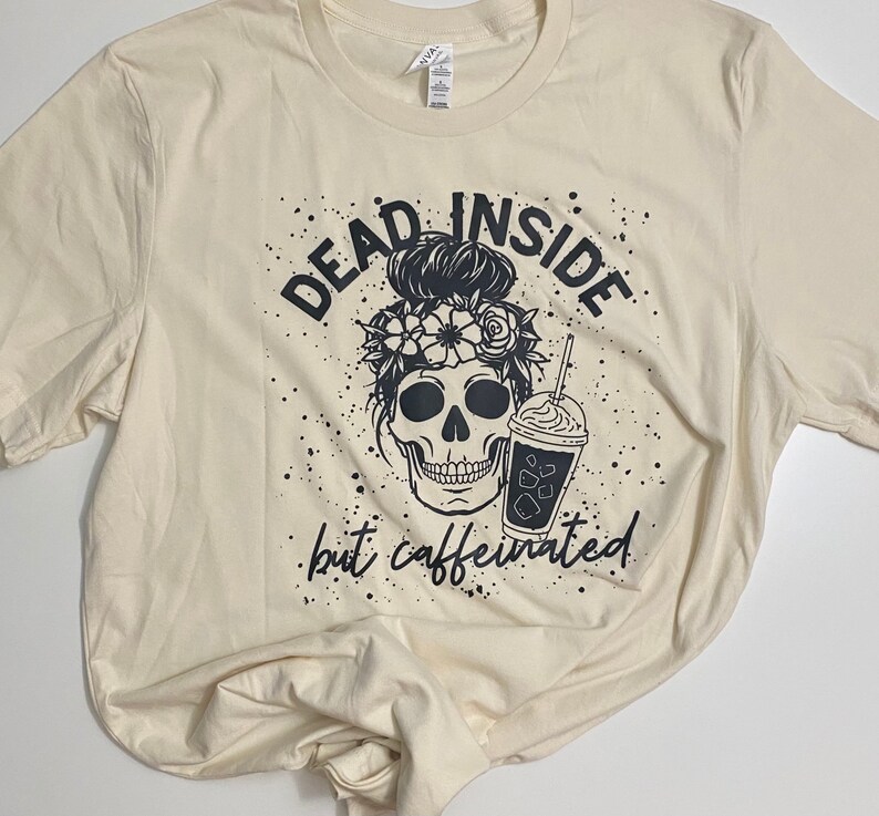 May include: A beige t-shirt with a black graphic design of a skull with flowers in its hair holding a iced coffee. The text "Dead Inside but Caffeinated" is printed below the skull.