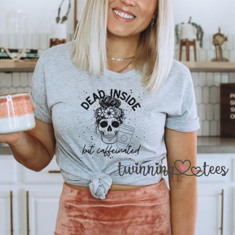 May include: Gray t-shirt with a black and white skull graphic. The skull is wearing a messy bun and holding a coffee cup. The text "Dead Inside but caffeinated" is printed below the skull. The text "twinnin' tees" is printed below the graphic.