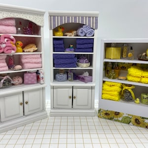 May include: Three miniature white wooden bathroom cabinets filled with tiny accessories. The left cabinet has pink towels and a yellow duck. The middle cabinet features purple towels and a yellow duck. The right cabinet displays yellow towels and sunflower decor.