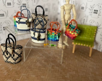 May include: A collection of miniature woven tote bags in various colors and patterns, including black and white stripes, rainbow stripes, and a brown and white pattern. The bags are displayed on a clear table with a wooden doll standing nearby.