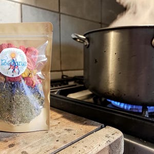 May include: A clear pouch filled with dried herbs and flowers, labeled "Balance." The pouch sits on a wooden surface next to a pot of boiling water on a gas stove. Steam rises from the pot, indicating the water is hot.