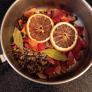 May include: A stainless steel pot filled with dried ingredients. Two dried orange slices are centered, surrounded by red rose petals, green bay leaves, and other dried herbs and spices. The pot sits on a black stovetop.