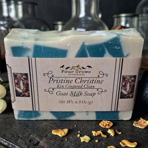 May include: A bar of goat milk soap with a blue and white design, wrapped in a label that reads "Pristine Christine" and "Goat Milk Soap." The soap is 4.3 oz. A black crow figurine and decorative elements are in the background.