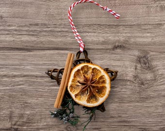 May include: A rustic Christmas ornament in the shape of a star, made of woven twigs. It features a dried orange slice, a cinnamon stick, and sprigs of greenery. A red and white twine loop is attached for hanging. The ornament is set against a wooden background.