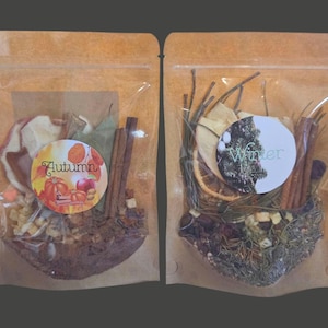 May include: Two clear cellophane bags filled with dried botanicals. One bag has a label that says "Autumn" with images of pumpkins and apples. The other bag has a label that says "Winter" with a winter scene. Cinnamon sticks and dried fruit are visible.