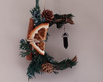May include: A crescent-shaped wall hanging made of green pine branches, brown pinecones, a dried orange slice, a cinnamon stick, and small blue berries. A black crystal pendant hangs from the center, and the piece is suspended by twine.