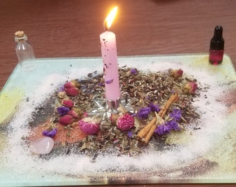 May include: A lit pink candle in a silver holder sits on a decorative glass plate surrounded by dried flowers, herbs, and a small bottle. A small glass bottle with a cork and a small bottle with a dropper are also present. The scene evokes a mystical or spiritual theme.