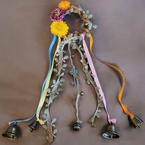 May include: A decorative wreath with a circular grapevine base, adorned with colorful ribbons, faux greenery, and small bronze bells. The wreath features a cluster of dried flowers in yellow, orange, and purple hues, and a decorative key charm.
