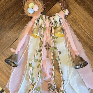 May include: A decorative wreath with a woven twig base, adorned with small pink roses and a nest holding three speckled eggs. Pink and white ribbons hang below, accented with gold bells, a key charm, and faux greenery.