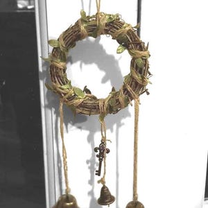 May include: A decorative wreath made of woven twigs, adorned with small green leaves and twine. Three brass bells and a small key charm hang from the wreath, creating a rustic and charming aesthetic.