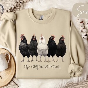 Puede incluir: Una sudadera beige con un gráfico de cinco pollos, cuatro negros y uno blanco, con el texto "My Crew is Fowl" impreso debajo. La sudadera está sobre una superficie blanca con una bandeja de madera decorativa y una taza blanca.