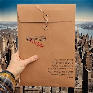 May include: A brown paper envelope with a string and two metal buttons. The envelope has a red stamp that says "CLASSIFIED" and the text "MOON RISING MYSTERIES PRESENTS: THE MANHATTAN MYSTERY GAME". The text "'AN INTERACTIVE. MYSTERY SOLVING, CASE FILE DELVING. ESCAPE ROOM-ISH (IT'S NOT AN ESCAPE ROOM) PROBING, HANDMADE GAME'" is printed on the envelope.