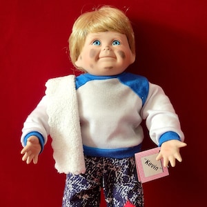 May include: A vintage doll named "Kevin" with blonde hair, blue eyes, and a painted face. The doll wears a white and blue raglan shirt, patterned pants, and a white towel. A red patch is sewn on the pants. The doll is barefoot.