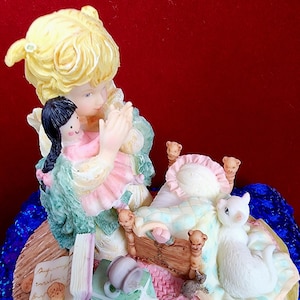 May include: A porcelain figurine of a girl kneeling in prayer with a doll in her arms. She is wearing a green and white dress and has blonde hair. The figurine is set on a wooden base with a bed, a cat, and a mouse.