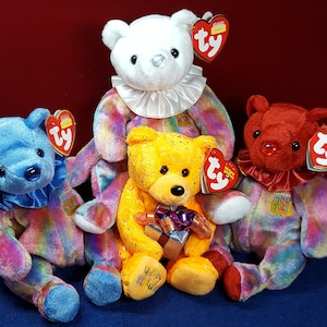 May include: Four colourful plush beanie baby bears with birthday themed designs. The bears are sitting on a blue surface. The bears are wearing colourful hats and have a rainbow pattern on their bodies. The bears have a Ty tag attached to their ears.